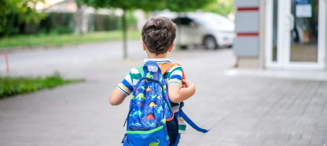 boy with blue backpast and striped shirt walking alone towards trees and building