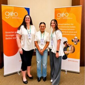 Anna, Duey, and Angela standing in white shirts in front of two GEO conference banners